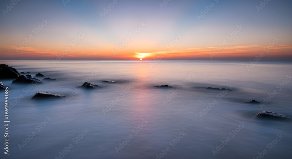 Fototapeta premium Serene ocean sunset with soft, blurred waves gently washing over dark, wet rocks on the shore, creating a tranquil and peaceful atmosphere
