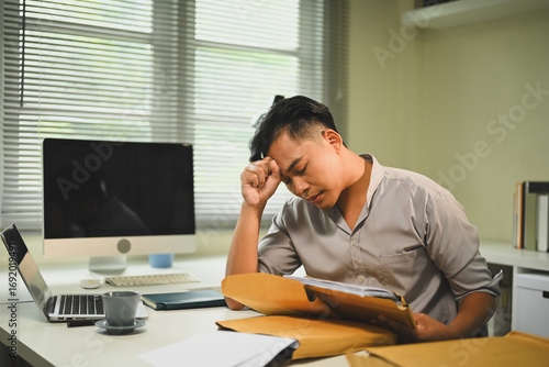 Stressed office worker sitting at desk with paperwork, holding his head while reviewing documents