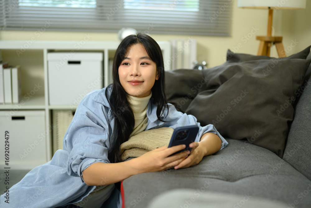 Obraz premium Young woman relaxing on sofa holding smartphone, smiling thoughtfully