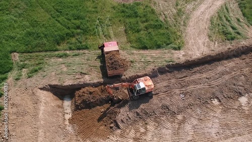 Aerial footage of an excavator loading soil into a dump truck on a dirt construction site.