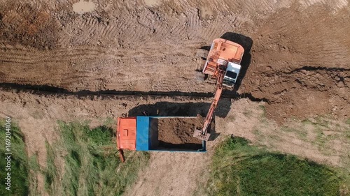 Aerial footage of an excavator loading soil into a dump truck on a dirt construction site.