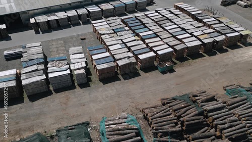 A drone captures stacks of cut timber and wooden planks stored outdoors in an industrial lumber yard. A systematic arrangement shows large-scale wood processing in progress.