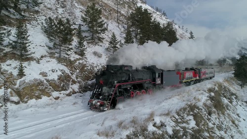 From above authentic vintage steam train journey through snowy mountain railway in winter sunshine. White steam rising from locomotive chimney, traveling to remote destinations, unique tour adventure