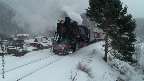 Aerial approach historical steam train stopped at Listvyanka station on Lake Baikal during winter snowstorm. Circum-Baikal railway with authentic village background, heavy snowfall, steam from chimney
