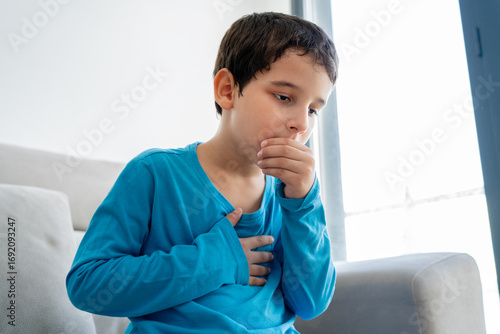 Little boy sitting on sofa at home and holding hand on chest. Male having asthma attack, difficulties with breathe, feeling severe pain or dyspnea