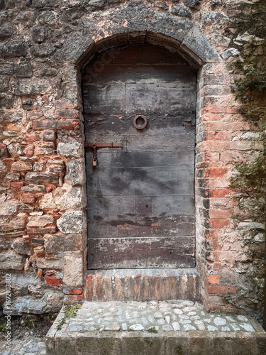 A rustic, weathered wooden door with an iron ring and padlock is set within an arched entryway of an old, crumbling brick and stone wall.