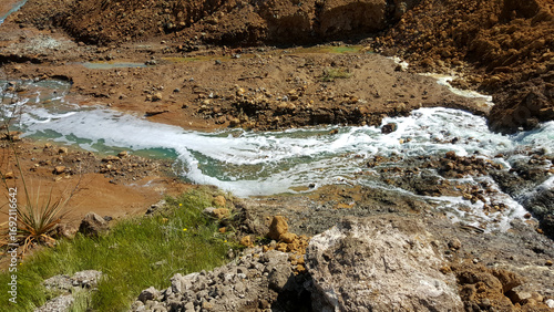 A polluted stream with rusty, orange-colored water flowing through a barren landscape, illustrating the severe environmental impact of mining waste and toxic runoff.