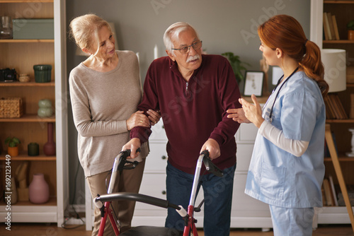 In a bright living room, a home health aide assists an elderly man using a walker. His wife supports his arm as the caregiver provides instructions or encouragement.