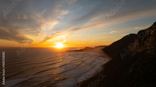 Aerial view of the golden sun kissing the ocean's edge, casting a warm glow over the rugged coastline, Cape Town, Western Cape, South Africa.