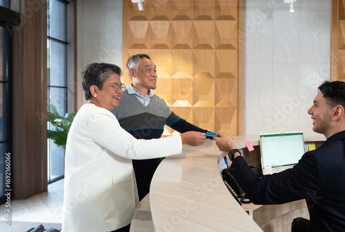 Senior asian couple interact with a hotel receptionist while checking in to their room. The friendly staff member assist them at the sleek front desk in a modern setting.