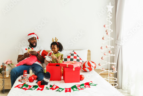 Happy father and child wearing festive accessories open Christmas gifts on a cozy bed with decorations and a minimalist tree in a bright home interior.