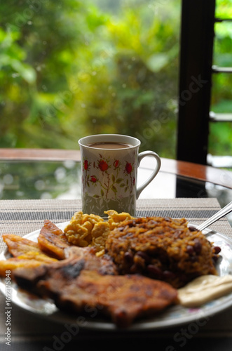 A top-down vertical view of a plate of delicious gallo pinto and pork chop with plantain, scrambled egg, and custard on a wooden table, a typical Costa Rican dish.