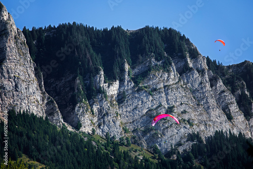 Parapentistes frôlant les falaises rocheuses et abruptes, garnies de sapins, sous le soleil et le ciel bleu d'été, dans les Alpes.