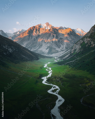 Aerial view of a winding river snaking through a vibrant green valley, framed by rugged mountains kissed with golden light, Karakol, Issyk-Kul Region, Kyrgyzstan.