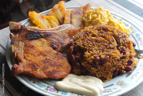 A top view of a plate of delicious Gallo pinto and pork chop with plantain, scrambled egg, and custard on a wooden table, a typical dish of Costa Rica.