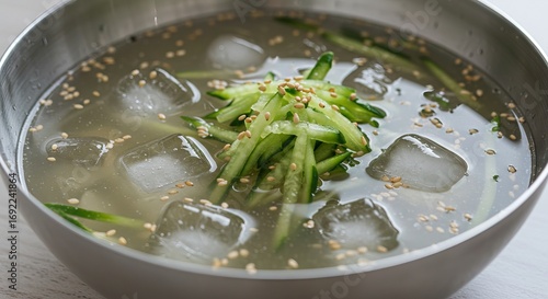 Cold noodle soup in a metal bowl with ice cubes cucumber strips and sesame seeds