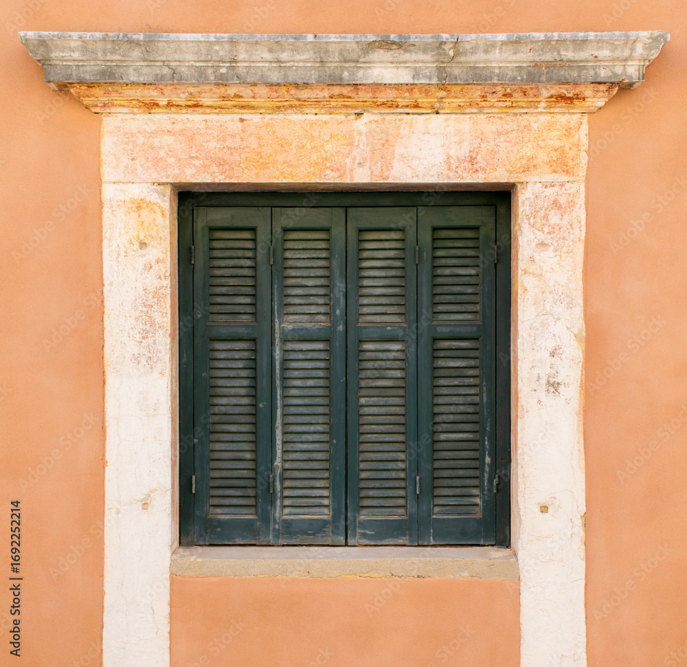 Fototapeta premium Dark green wooden shutters closed on a weathered stone-framed window set in a textured orange wall