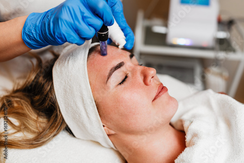 Beautician wearing blue gloves uses a microneedling pen to apply serum to forehead of client with closed eyes lying on a treatment table during a beauty spa procedure. Modern anti-age procedure.