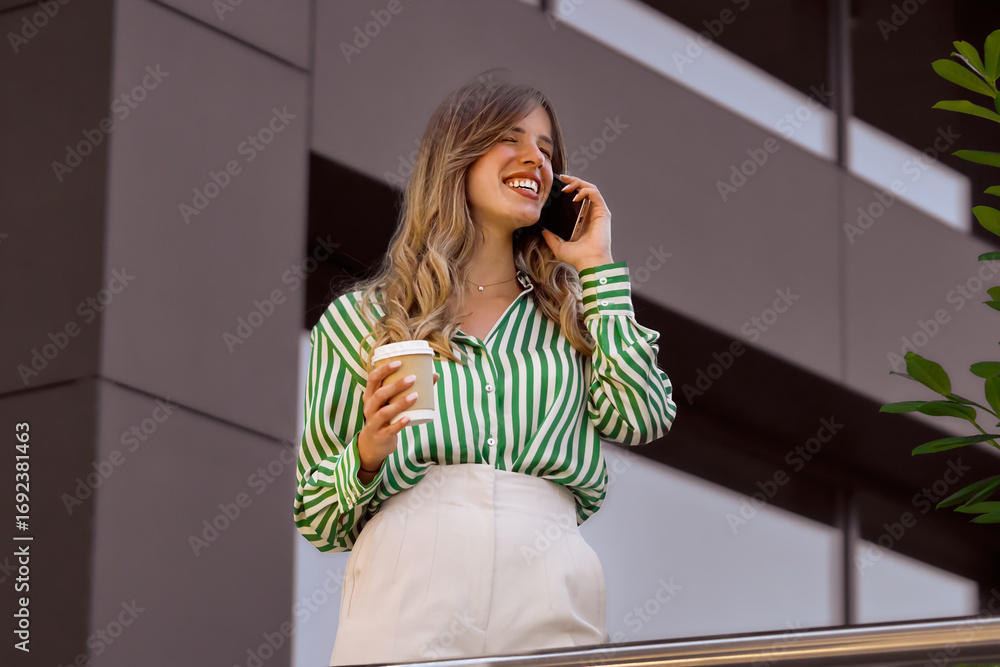 Fototapeta premium Confident woman with striped shirt enjoys a phone call while holding a coffee cup outdoors, exuding positivity and a professional vibe in an urban environment during daytime.