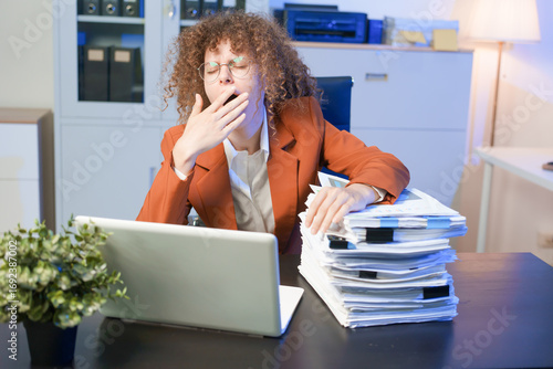 Tired young woman working late at the office, looking sleepy, using smartphone, wanting to relax, working overtime, concept of overtime deadline.