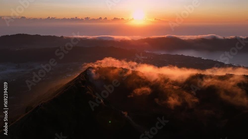 A golden sunrise cast light over Mount Batur in Bali, Indonesia, with hikers gathered at the summit to witness the mist veiling volcanic peak and glowing clouds.