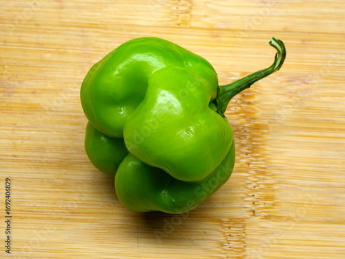 A photograph of a unique and interestingly shaped organic green peter pepper with a wrinkled texture and long curved stem on a wooden cutting board surface