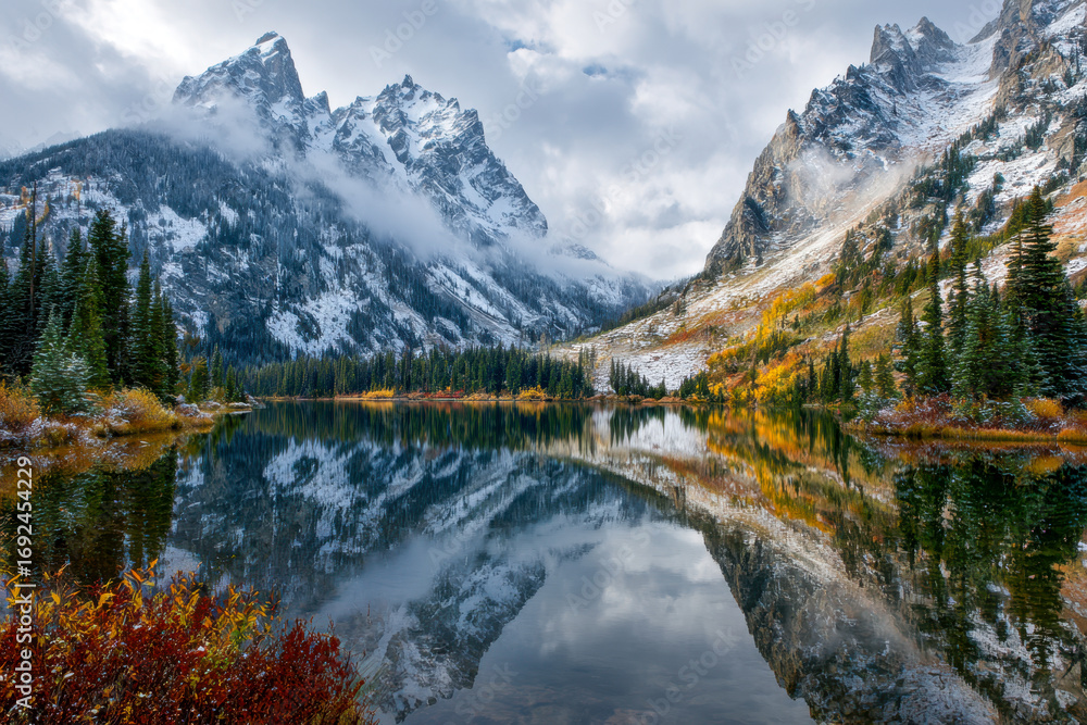 Naklejka premium Tranquil lake reflecting snow-covered rugged mountain peaks and colorful autumn foliage under a cloudy sky in a serene wilderness landscape