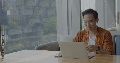 Senior Asian businessman wearing headset celebrates success during online meeting in modern co-working space. Remote work and business communication concept	
