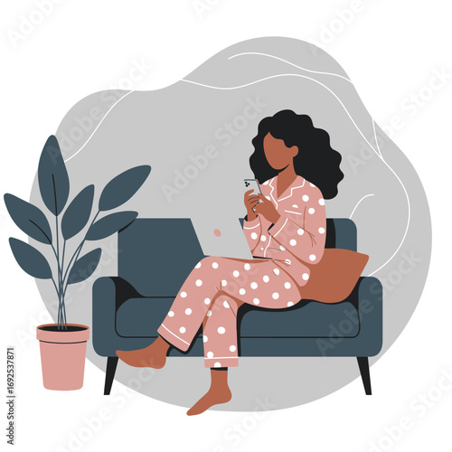 African American woman in pajamas working from home on a laptop and phone, embracing remote work and digital convenience from her cozy sofa.