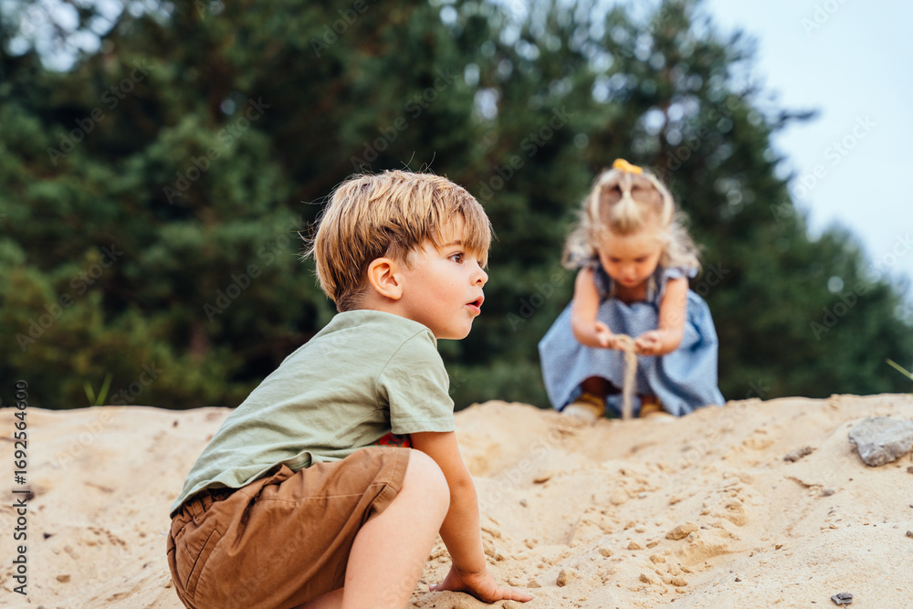 Fototapeta premium Boy playing on sand hill with sister in background during summer day