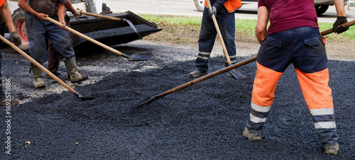Foto Road crew in hi-vis orange spreads and levels fresh asphalt with shovels on a city street; summer heat, hard manual work and urban upkeep in progress