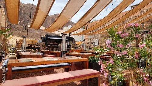 Outdoor cafe in AlUla, Saudi Arabia, with wooden tables and benches under beige fabric canopies, offering shade. The setting includes potted plants with pink flowers and a food truck backdrop.