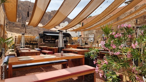 Outdoor cafe in AlUla, Saudi Arabia, with wooden tables and benches under beige fabric canopies, offering shade. The setting includes potted plants with pink flowers and a food truck backdrop.