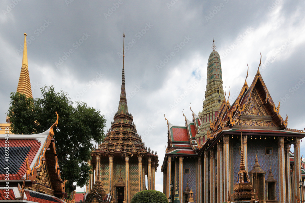 Fototapeta premium Temple roofs at Grand Palace, Bangkok - A wide shot of the ornate and majestic rooftops of the Grand Palace, showcasing traditional Thai architectural design under a cloudy sky.