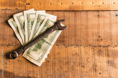 Leather work gloves holding a rusty wrench and American dollars on a rustic wooden work table