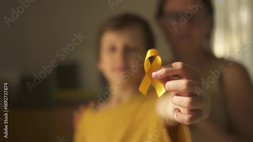 Adolescent boy holding yellow childhood cancer awareness ribbon while his mother provides comforting shoulder embrace. United in support.