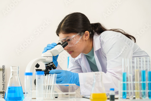 Asian teenage girl with long hair tied back wears lab coat and protective glasses, working attentively at microscope in science lab, exploring fascinating details of chemical and biological samples.