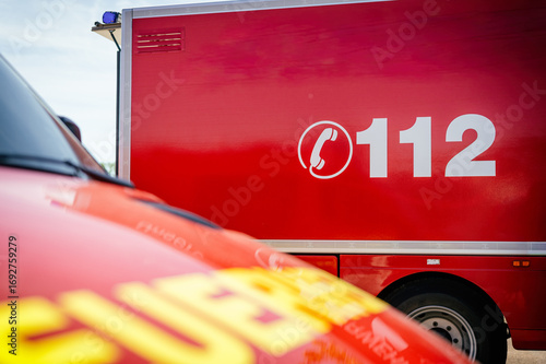 Rotes Feuerwehrfahrzeug mit der Notrufnummer 112, im Vordergrund ein weiteres Einsatzfahrzeug unscharf. Symbolisiert Notruf, Einsatzkräfte, Rettungsdienst und öffentliche Sicherheit.