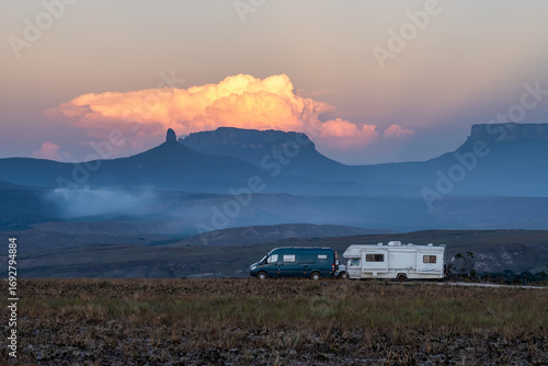 Two overlanding vehicles parked in the vast Gran Sabana, gazing at majestic tepui mountains shrouded in sunset clouds. A scene of travel and van life adventure in Venezuela