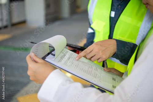 Inspection sheet being reviewed by two workers wearing safety vest in industrial environment showing detailed data and checklist for quality control and safety compliance