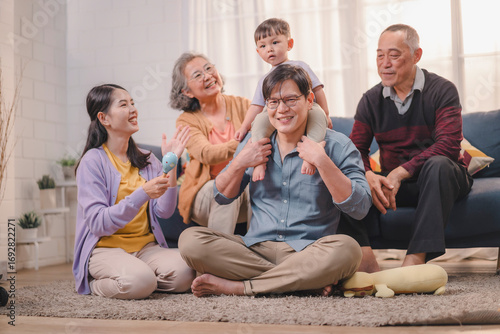 Happy Asian multigenerational family spending time together at home. Father carries toddler on shoulders while mother and grandparents laugh and play, capturing warmth and family bonding.