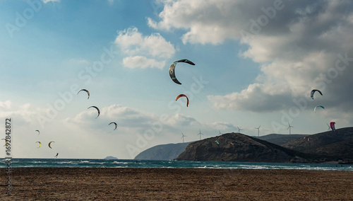 Kitesurfers riding waves with colorful kites flying above beach and mountains in the background.