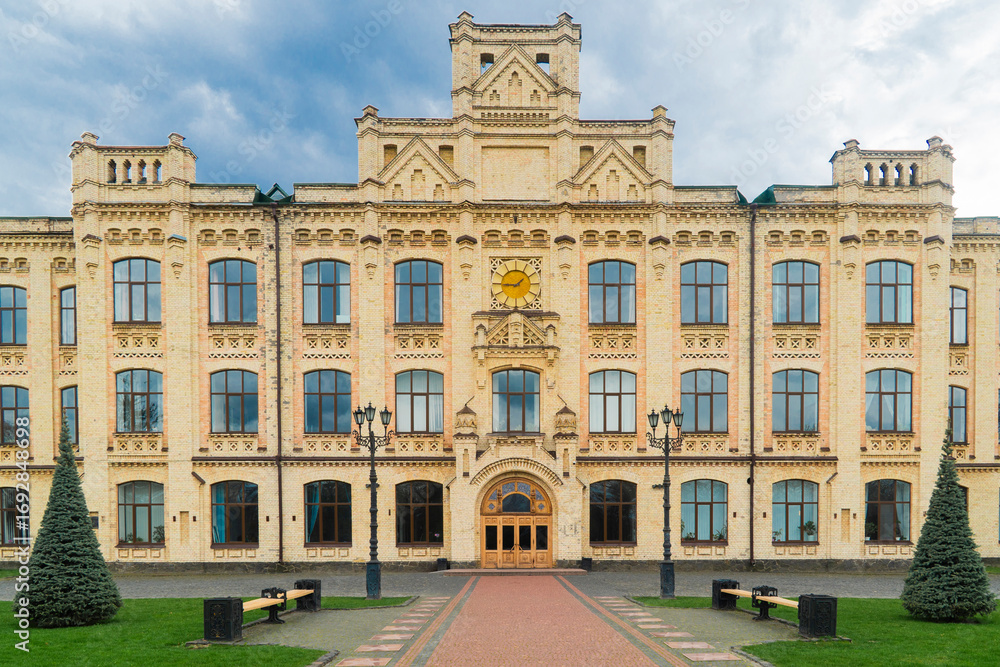 Fototapeta premium Kyiv. Kyiv Polytechnic Institute. Large, yellow brick building with detailed architectural features, numerous windows, and surrounding green lawns and trees on a cloudy day