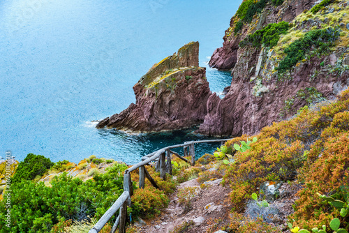 Breathtaking view of the turquoise Mediterranean Sea meeting the unique red rocks of Cala Zurletto in Capraia Island, with a wooden fence guiding the way along the cliffside vegetation