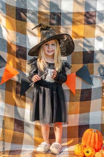 A little girl in a black dress and a witch hat dressed up for Halloween, flags on an orange plaid and pumpkins