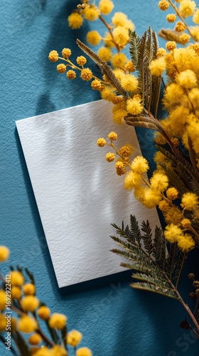 Creative Arrangement of Yellow Flowers and Blank Paper Against Blue Wall Back...