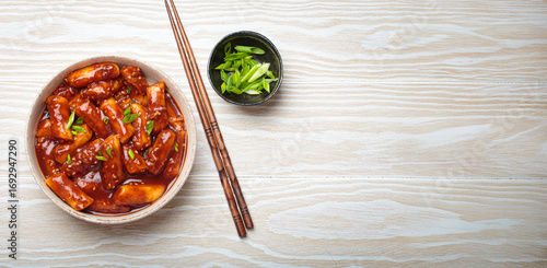 Stir fried rice cakes tteokbokki simmered in spicy sauce, garnished with green onions and sesame seeds, served on a white wooden background with chopsticks top view. Popular dish of Korea, copy space