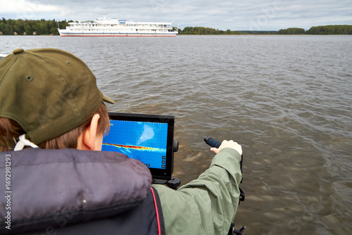 Fisherman steering boat while looking at fishfinder screen on a lake. Modern angling technology with echo sounder and scenic river view. Fishing, navigation, and outdoor recreation concept