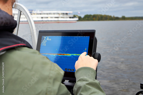Fisherman steering boat while looking at fishfinder screen on a lake. Modern angling technology with echo sounder and scenic river view. Fishing, navigation, and outdoor recreation concept