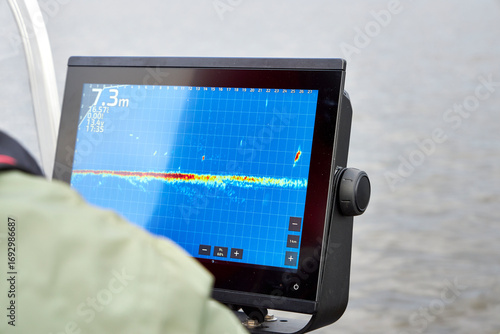 Fisherman steering boat while looking at fishfinder screen on a lake. Modern angling technology with echo sounder and scenic river view. Fishing, navigation, and outdoor recreation concept